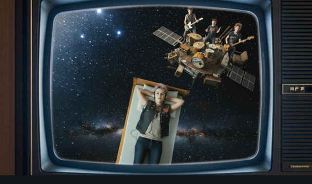 Still aus Trailer zum Song zu Mauerpogo von Sonja M. Schultz. Abgebildet wird ein Mädchen, das auf einem Bett liegt, das im Universum fliegt, dahinter eine spielende Band