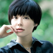 Porträtfoto Yukiko Motoya: Die Außenaufnahme zeigt die Autorin vor grünem Hintergrund. Sie blickt direkt in die Kamera und hat eine Hand, wie abwartend, an die Wange gelegt. Sie trägt die fast schwarzen Haare in einem Topfschnitt, eine dunkle Hemdbluse und dezenten Silberschmuck.
