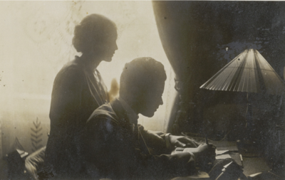  Schwarz-weiss Foto von Netty Reiling und Rodi. Er sitzt am Schreibtisch und schreibt, sie steht hinter ihm, beide im Profil