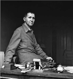 Schwar-weiss-Fotografie von Fred Stein. Abgebildet wird ein Mann, dr auf einem Tisch sitzt, auf dem Tisch befindet sich eine Schreibmaschine.