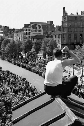 Schwarz-weiss-Fotografie von Fred Stein. Abgebildet wird ein junger Mann, der auf einem Dach sitzt und auf eine Menschenmenge auf der Straße schaut.