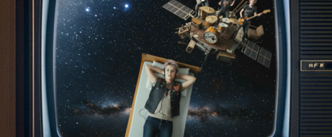 Still aus Trailer zum Song zu Mauerpogo von Sonja M. Schultz. Abgebildet wird ein Mädchen, das auf einem Bett liegt, das im Universum fliegt, dahinter eine spielende Band